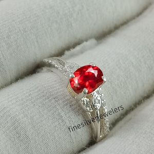 May include: A silver ring with a large red oval gemstone surrounded by smaller clear gemstones. The ring is set on a white background with the text "finesilverjewelers" visible.