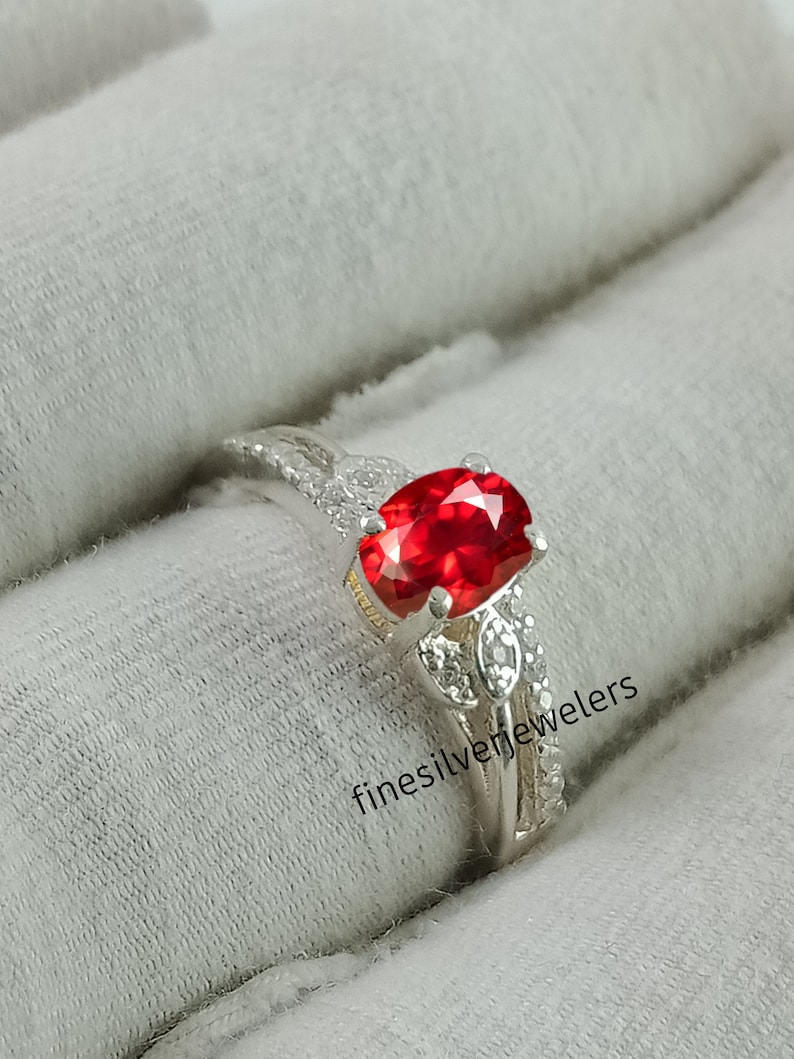May include: A silver ring with a large red oval gemstone surrounded by smaller clear gemstones. The ring is set on a white background with the text "finesilverjewelers" visible.