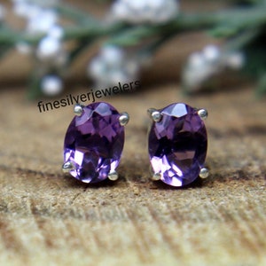 Puede incluir: Un par de pendientes de plata de ley con piedras preciosas de amatista de forma ovalada. Los pendientes están engastados en un diseño simple y elegante.