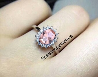 May include: A silver ring with a large oval pink gemstone surrounded by smaller clear gemstones. The ring is on a finger.