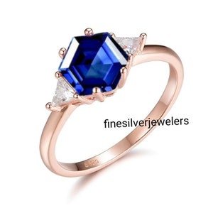 May include: A rose gold ring with a blue hexagonal sapphire gemstone and two smaller triangular diamonds. The ring is marked with "925" on the inside band. The text "finesilverjewelers" is visible below the ring.