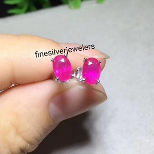 May include: A pair of silver earrings with oval-shaped pink ruby gemstones. The earrings are held in a hand.