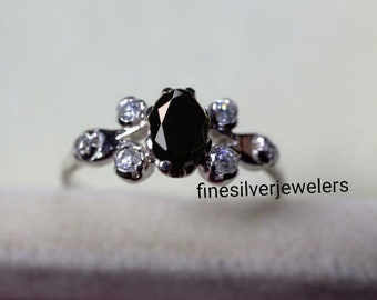 May include: A silver ring with a black oval gemstone surrounded by smaller white gemstones. The ring is on a white surface.  finesilverjewelers