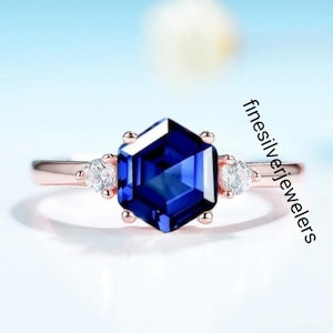 May include: A rose gold ring with a blue hexagonal gemstone and two smaller clear gemstones on the band. The ring is set on a white background.