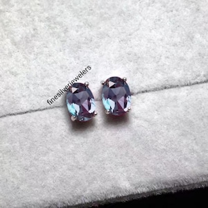 Könnte beinhalten: Ein Paar Ohrstecker mit oval geschliffenen Alexandriten in Silberfassungen. Die Steine zeigen ein Farbspiel mit Nuancen von tiefem Violett und Blaugrün. Der Text finesilverjewelers ist neben dem linken Ohrring deutlich zu erkennen.