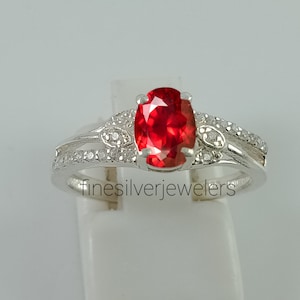 May include: A silver ring with a red oval gemstone and smaller clear gemstones set in the band. The ring is on a white display stand.  finesilverjewelers