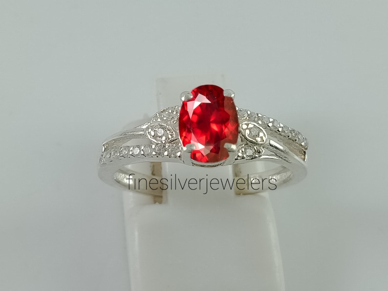 May include: A silver ring with a red oval gemstone and smaller clear gemstones set in the band. The ring is on a white display stand.  finesilverjewelers