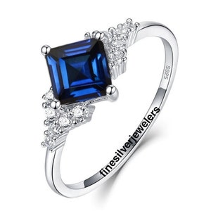 May include: A silver ring with a square blue sapphire gemstone and smaller clear gemstones. The ring is engraved with "finesilverjewelers" and "S925".