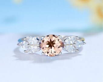 May include: A silver ring with a large, round, peach-colored gemstone surrounded by smaller, clear, marquise-shaped gemstones.