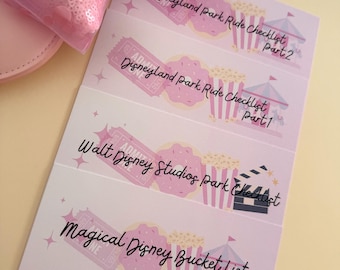 May include: Pink Disneyland and Disney Studios park checklists and a Magical Disney Bucket List, Paris Edition. The checklists feature popcorn, tickets, and a circus tent graphic. A pink sequined headband is in the background.