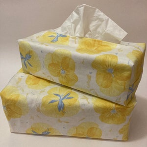 Könnte beinhalten: Zwei gelbe, florale Tissuebox-Bezüge mit weißem Hintergrund. Der obere Bezug hat eine weiße Tissuebox im Inneren.