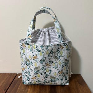 May include: A fabric tote bag featuring a light blue background with a pattern of green pears, white flowers, and small birds. The bag has two matching fabric handles and a grey and white striped drawstring closure at the top with white cords.