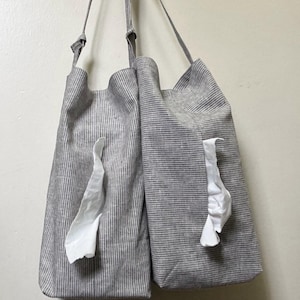 May include: Two grey and white striped fabric tote bags with long straps hang against a white wall. Each bag has a white tissue sticking out of a pocket. The bags are made of a soft, textured material.