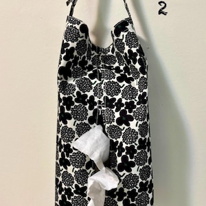 May include: A fabric tissue holder featuring a black and white floral design. The rectangular holder has a strap for hanging and a slot for dispensing tissues. White tissues are visible.