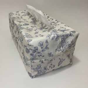 Könnte beinhalten: Ein rechteckiger Taschentuchbox-Bezug mit weißem Hintergrund und blauem Blumen- und Vogelmuster. Weiße Taschentücher ragen aus der oberen Öffnung heraus. Der Bezug ist aus Stoff und hat ein sauberes, fertiges Aussehen.