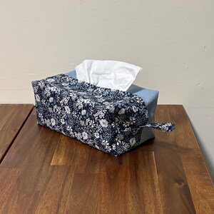 May include: A rectangular tissue box cover with a floral pattern in navy, white, and light blue. The box has a light blue base and a matching handle on one side. White tissues are visible at the top.