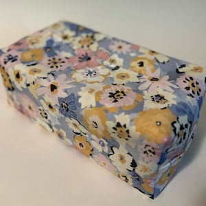 Könnte beinhalten: Eine rechteckige Schachtel, die mit einem Blumenmuster bedeckt ist, mit blauen, rosa, gelben und weißen Blumen auf einem hellgrauen Hintergrund.