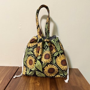 May include: A fabric drawstring bag featuring a repeating pattern of yellow sunflowers with brown centres and green leaves on a black background. The bag includes a single top handle and white cord drawstrings on both sides for closure.