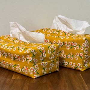 May include: Two rectangular tissue box covers. They are covered in a yellow floral fabric with white and orange daisy-like flowers. White tissues are pulled from the top of each box. The boxes are on a wooden surface.