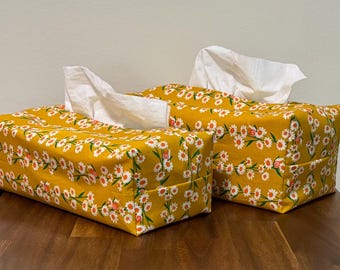 May include: Two rectangular tissue box covers. They are covered in a yellow floral fabric with white and orange daisy-like flowers. White tissues are pulled from the top of each box. The boxes are on a wooden surface.