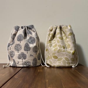 May include: Two drawstring bags. One bag features a pattern of grey trees and rabbits on a beige background. The other bag has a pattern of green leaves on a beige background. Both bags have white drawstrings.