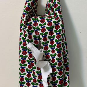 Könnte beinhalten: Ein Stoff-Taschentuchbox-Bezug mit einem sich wiederholenden Blumenmuster aus roten Rosen, grünen Blättern und blauen Akzenten auf weißem Grund. Rechteckige Form mit Schlitz zur Entnahme der Taschentücher und einem Griff.