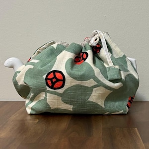 May include: A fabric tea cozy with a white ceramic spout. The cozy is patterned with green leaf shapes and red circular designs on a cream background. The tea cozy is sitting on a wooden surface.
