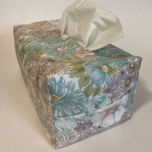 Könnte beinhalten: Ein floral bedruckter Taschentuchbox-Bezug mit einem hellblauen, grünen und braunen Muster. Der Bezug ist aus Stoff und hat eine quadratische Form.