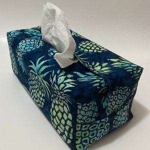 Peut inclure: Un couvre-boîte à mouchoirs bleu et vert avec un motif d'ananas. Le couvre-boîte est en tissu et possède une fente pour sortir les mouchoirs.
