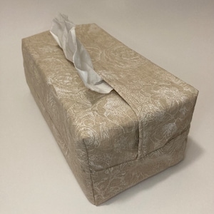 Könnte beinhalten: Ein beigefarbener Tissuebox-Bezug aus Leinen mit einem weißen Blumenmuster. Der Bezug hat einen Schlitz für Taschentücher und eine Klappe, um sie an Ort und Stelle zu halten.