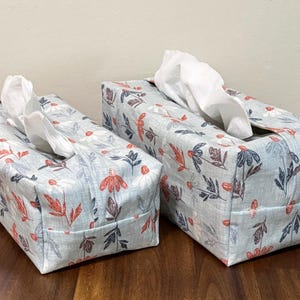 Peut inclure: Deux boîtes à mouchoirs rectangulaires avec des motifs floraux. Les housses sont bleu clair avec des motifs floraux rouges et marine. Des mouchoirs blancs sont visibles par les ouvertures supérieures. Les boîtes sont sur une surface en bois.