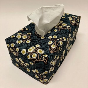 Könnte beinhalten: Ein schwarzer Taschentuchbox-Bezug mit einem Blumenmuster mit weißen Blüten und braunen Stielen auf dunkelblauem Hintergrund.