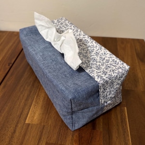Könnte beinhalten: Ein rechteckiger Taschentuchbox-Bezug aus blauem Denim-Stoff mit floral gemusterter Oberseite. Weiße Taschentücher ragen aus der Öffnung heraus. Das Blumenmuster ist weiß mit kleinen marineblauen Blumen. Die Box steht auf einer Holzoberfläche.