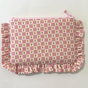 May include: A pink and white checkered zippered pouch with a floral pattern. The pouch has a ruffled edge and a pink zip pull. The pattern includes small yellow flowers.