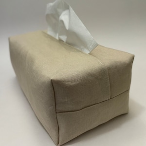 May include: A beige linen tissue box cover with a white tissue box inside. The cover has a simple design and is made of a soft, natural fabric.
