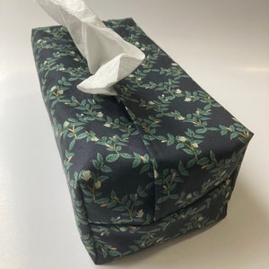 Könnte beinhalten: Ein schwarzer Taschentuchbox-Bezug mit einem floralen Muster aus grünen Blättern und weißen Blüten. Der Bezug hat eine weiße Taschentuchbox im Inneren.