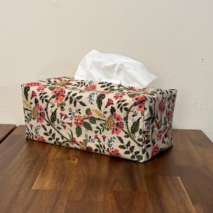 May include: A rectangular tissue box cover with a floral pattern in pink, red, yellow, and green on a cream background. White tissues are visible at the top. The box is on a wooden surface.