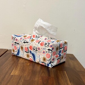 May include: A rectangular tissue box cover with a floral pattern in pink, blue, and gold on a white background. White tissues protrude from the top. The box rests on a dark wooden surface.