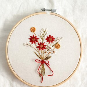 Może przedstawiać: Haftowany bukiet kwiatów w drewnianym tamborku. Bukiet zawiera czerwone, białe i żółte kwiaty związane czerwoną wstążką. Haft znajduje się na białym tle materiału.