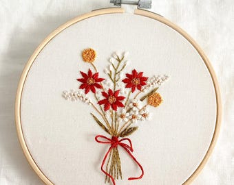 May include: Embroidered floral bouquet in a wooden hoop. The bouquet features red, white, and yellow flowers tied with a red ribbon. The embroidery is on a white fabric background.