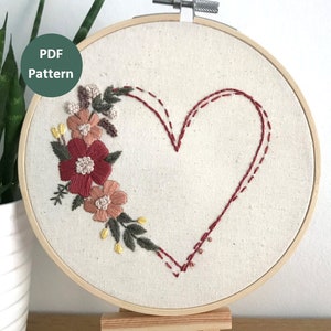 May include: Embroidered hoop art featuring a red heart with a floral design in shades of pink, red, and green. The hoop is made of wood and is displayed on a wooden easel.