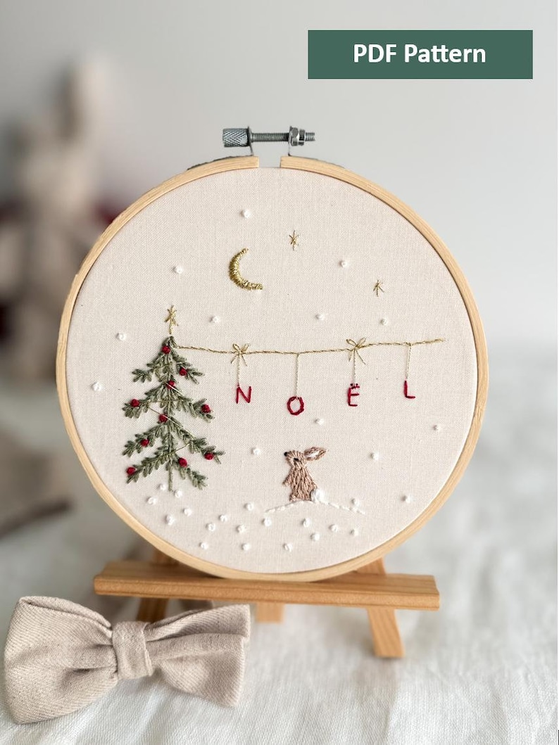 Może przedstawiać: Haftowany obręcz z motywem choinki, kr&oacute;lika i napisem "NO&Euml;L" w śnieżnym krajobrazie. Obręcz stoi na drewnianym stojaku.