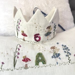 May include: A cream fabric crown with embroidered fairy tale details. The crown has a sequined number 6, fairies, mushrooms, and floral patterns. The letter "A" is also embroidered. A blue ribbon is attached. The design is whimsical.