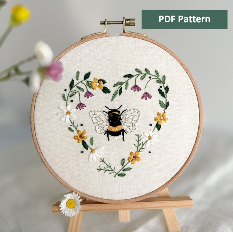 Peut inclure: Art de broderie sur un cercle repr&eacute;sentant une abeille dans une couronne florale en forme de c&oelig;ur. L'abeille est noire et jaune avec des ailes blanches. Les fleurs sont blanches, jaunes et violettes.