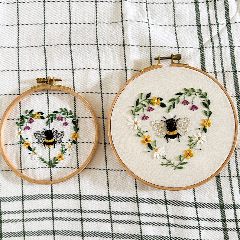 Peut inclure: Deux cerceaux &agrave; broder avec un tissu blanc et un motif en forme de c&oelig;ur repr&eacute;sentant une abeille et des fleurs. Le plus petit cerceau est &agrave; gauche et le plus grand &agrave; droite. Les deux cerceaux sont sur une nappe &agrave; carreaux blancs et verts.