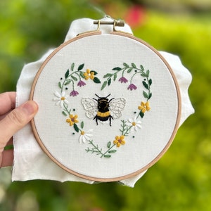 Peut inclure: Art de broderie sur un cercle repr&eacute;sentant un bourdon au centre d'un motif floral en forme de c&oelig;ur. Les fleurs sont jaunes, blanches et violettes.
