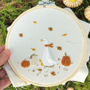 以下が含まれることがあります： 木製の刺繍枠に、オレンジ色のリボンをつけた白いガチョウ、カボチャ、秋の葉が刺繍されています。デザインはクリーム色の生地に刺繍されています。カボチャはオレンジと白です。