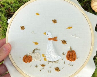 May include: Embroidery hoop featuring a white goose with an orange bow, two pumpkins, and autumn leaves. The design is stitched on a cream-colored fabric, set in a wooden hoop. The pumpkins are orange and white.