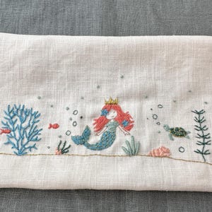 May include: Embroidered white fabric featuring an underwater scene. A mermaid with red hair and a gold crown is surrounded by coral, fish, bubbles, and a sea turtle. The design uses blue, red, and green thread.