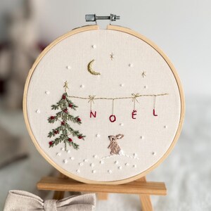 Może przedstawiać: Haftowany obręcz z zielonym drzewem bożonarodzeniowym, białym kr&oacute;likiem i napisem "NOEL" czerwonymi literami. Obręcz znajduje się na drewnianym sztalugach.